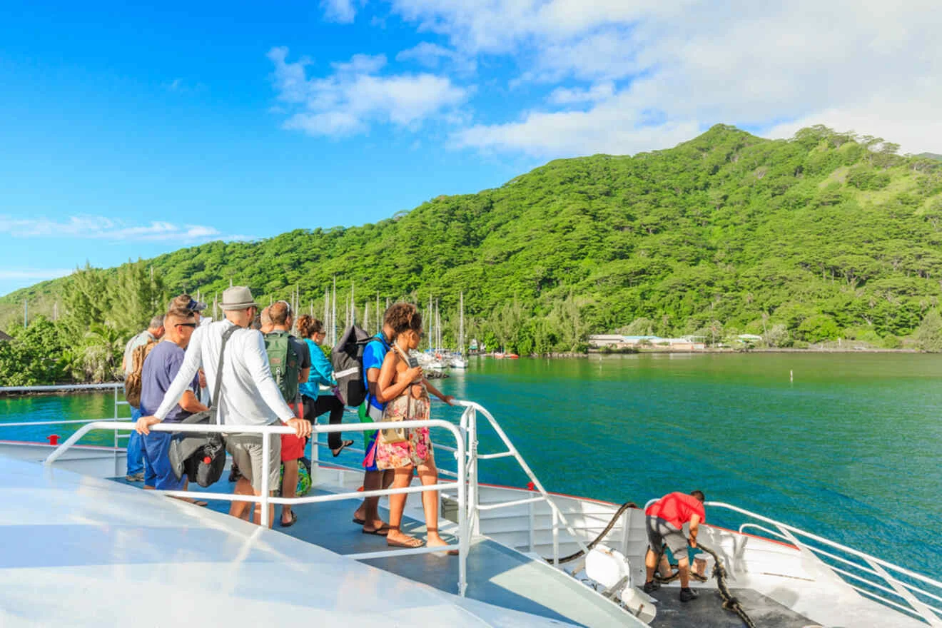 A group of people stands on a boat deck overlooking a lush green island and calm blue water on a sunny day. Some hold cameras and phones, while a crew member tends to ropes.