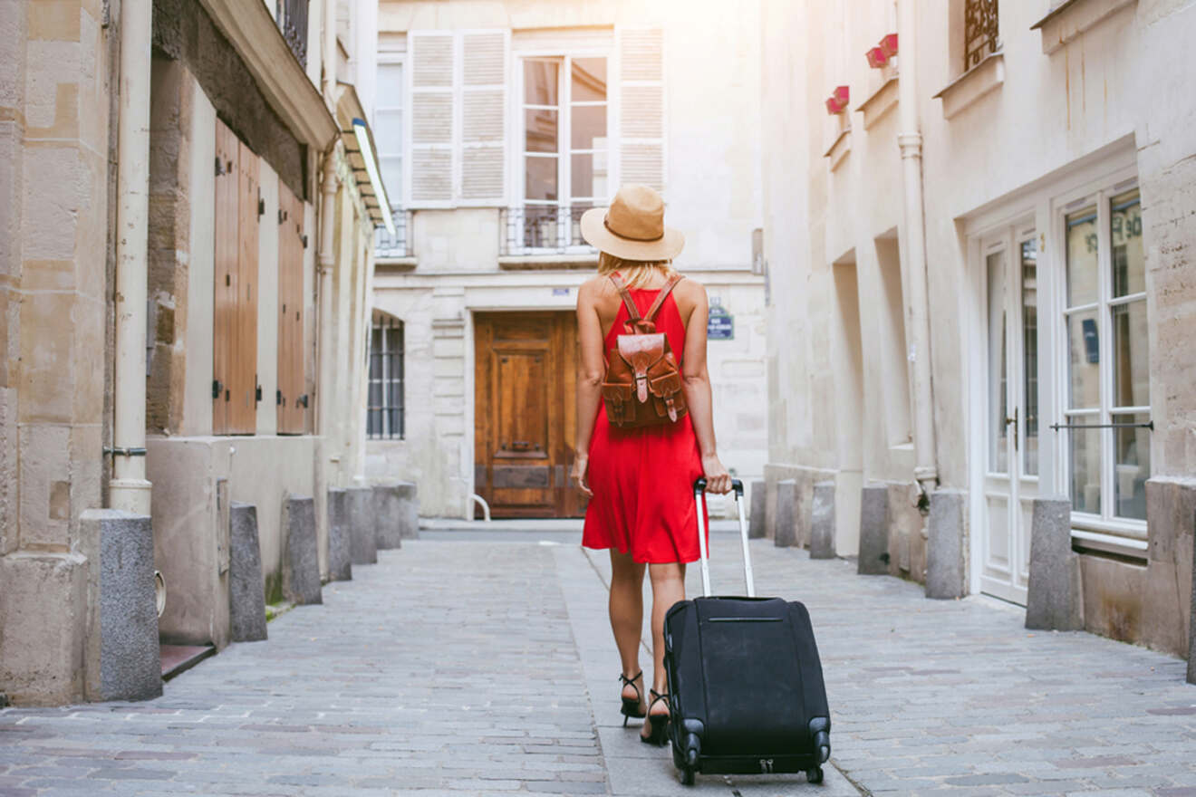 A woman in a red dress and hat walks down a narrow street with a rolling suitcase and a backpack.