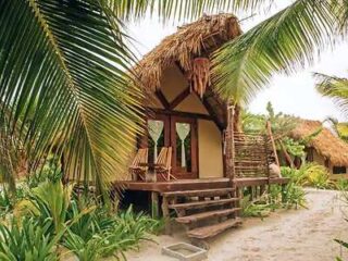 A small thatched-roof hut with a wooden porch and two chairs is surrounded by tropical plants and palm trees on a sandy surface.