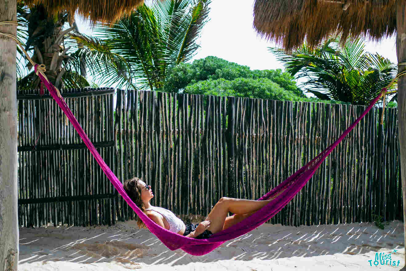 Yulia, the founder of this website,  is lying in a purple hammock under a thatched roof, with palm trees and a bamboo fence in the background.