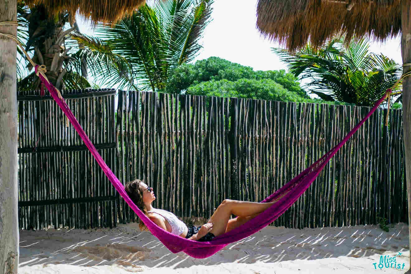 the author of the post relaxing on a purple hammock under a thatched roof on a sandy beach with palm trees and a bamboo fence in the background.