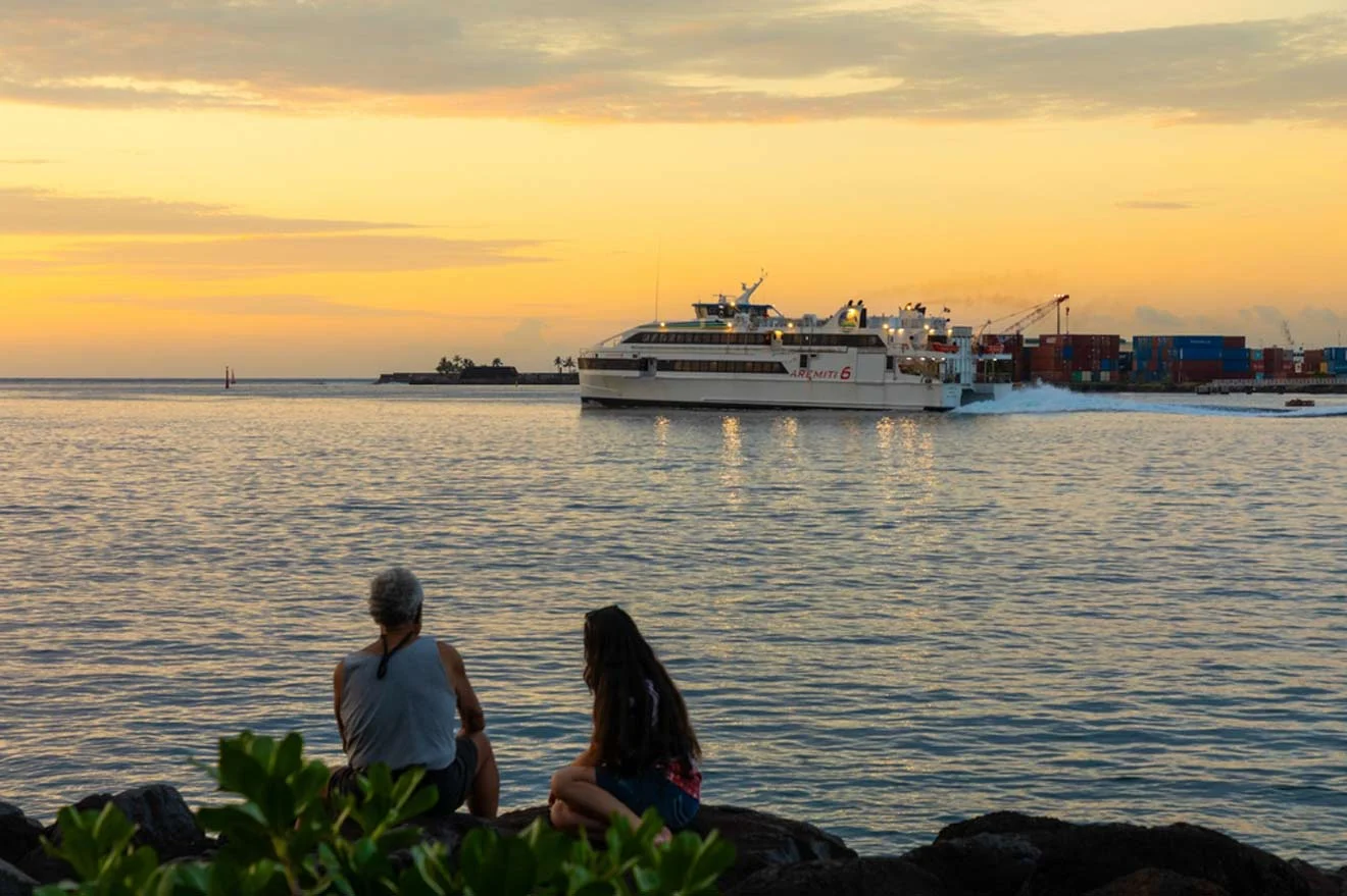 Two people sitting on rocks near the water's edge observing a large ferry boat at sunset.