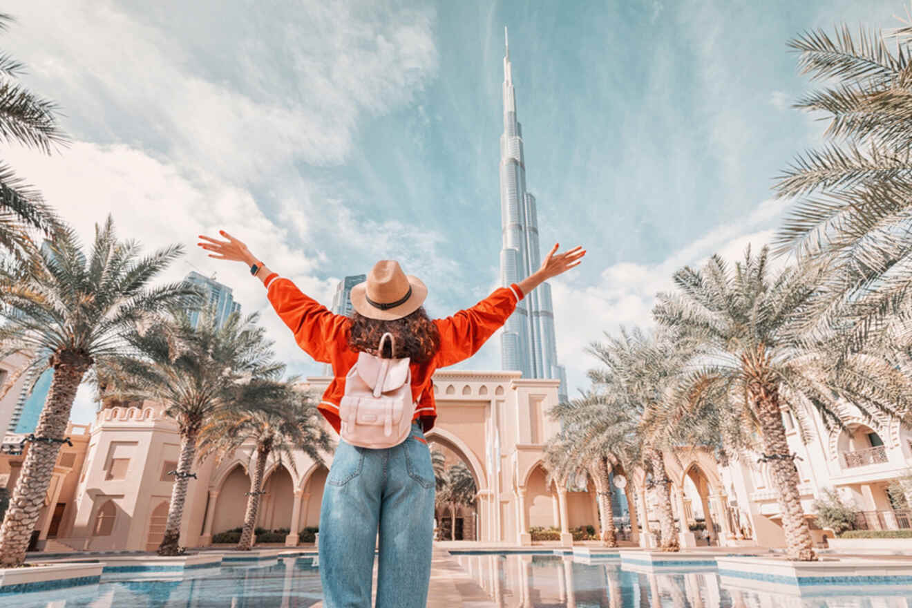 Person standing with arms raised, facing a tall skyscraper, surrounded by palm trees and architecture in a sunny setting.