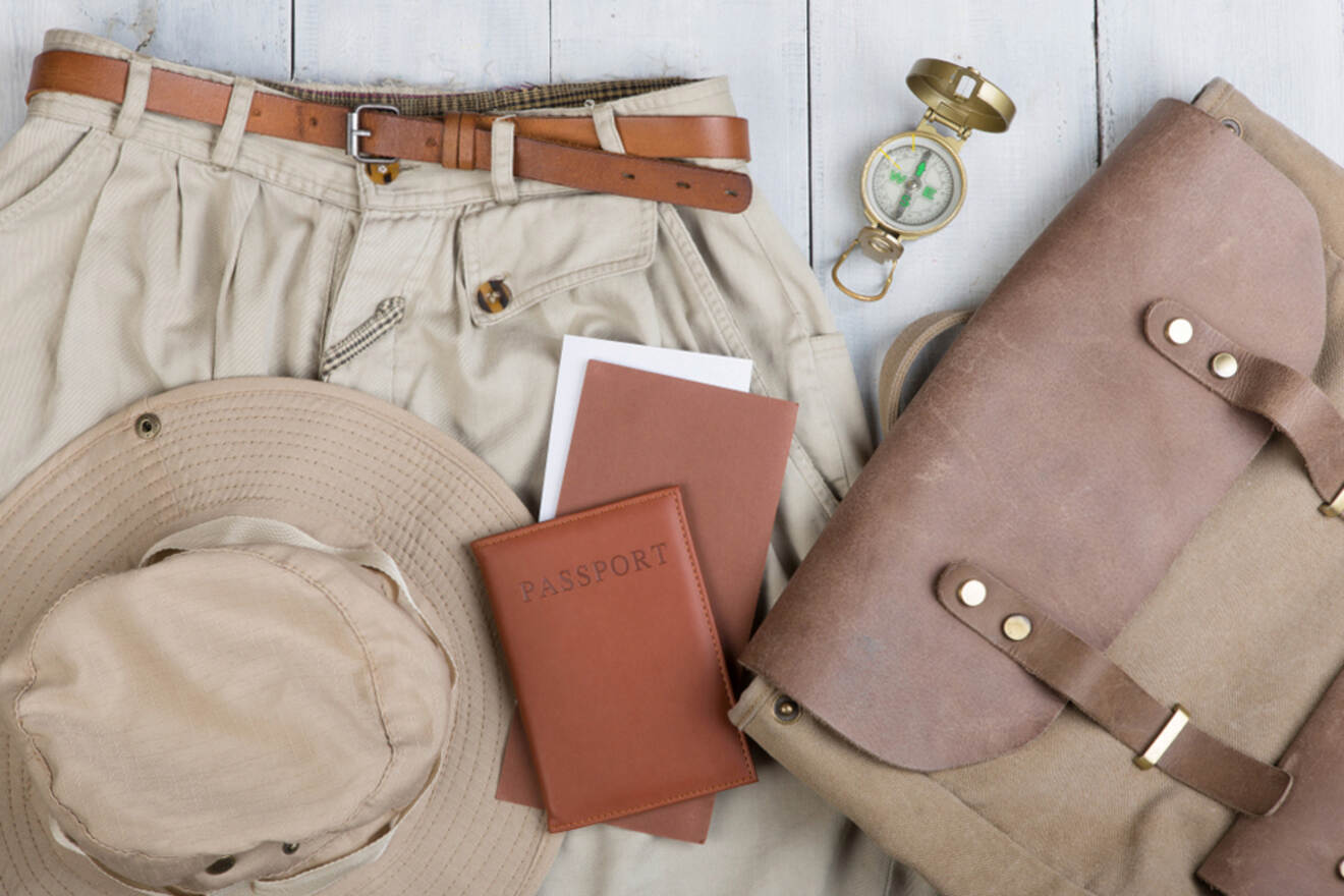 Assorted travel items including a brown leather bag, a khaki hat, a compass, a passport, a pair of beige pants with a brown belt, and two small notebooks, all laid out on a wooden surface.