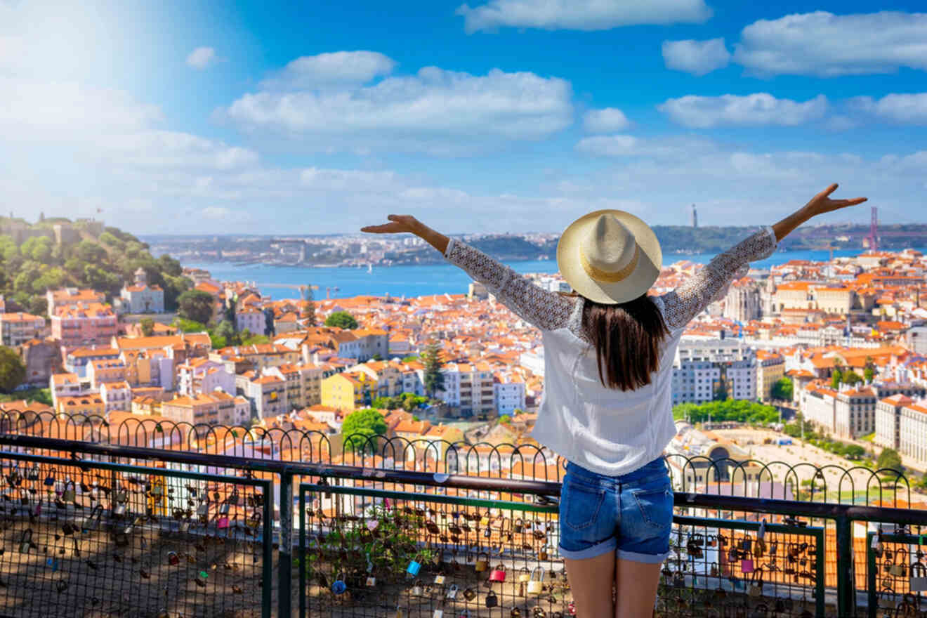 A woman in a hat and casual clothing stands with arms outstretched, facing a panoramic view of a cityscape with red-roofed buildings and a river in the background under a blue sky.
