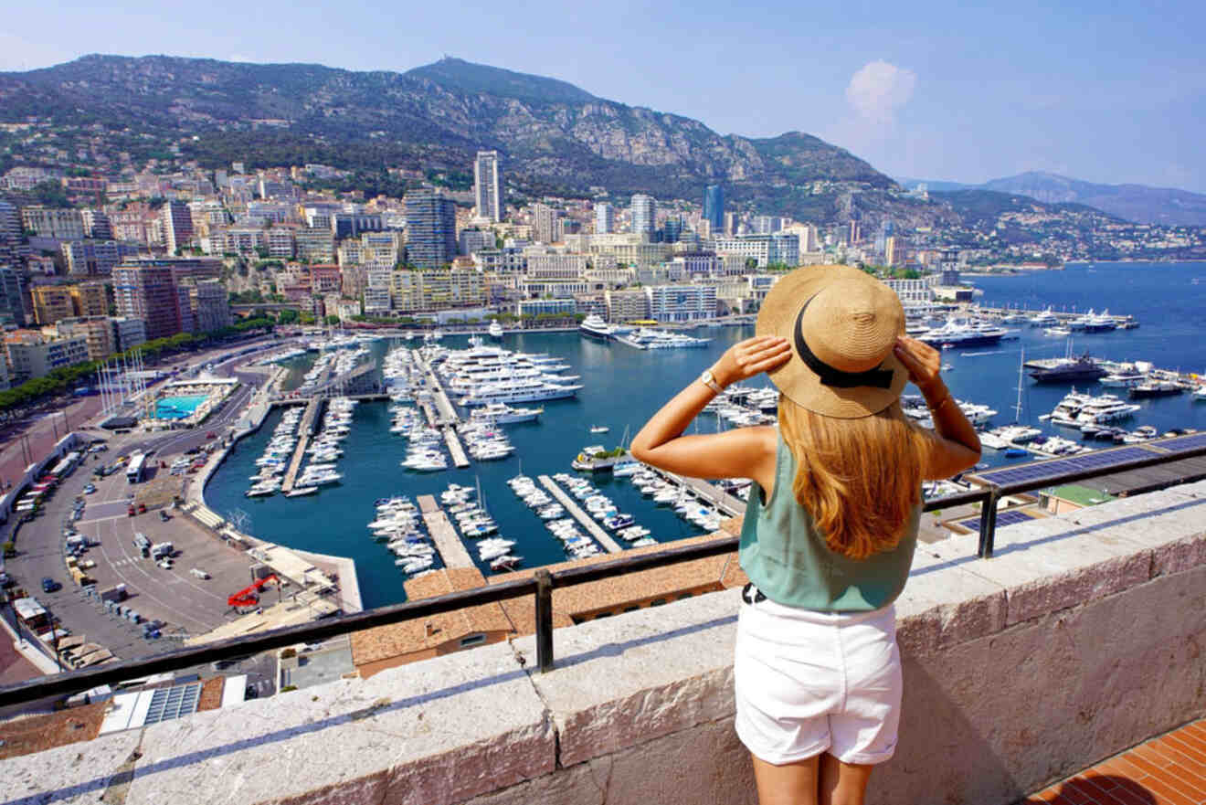 A woman in a sunhat and summer outfit overlooks a marina filled with yachts against a backdrop of the cityscape and mountains of Monaco.