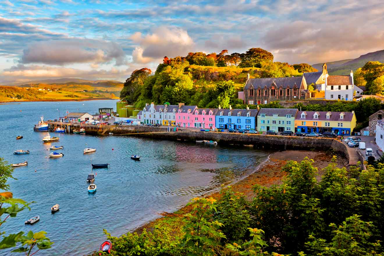 Colorful houses line the waterfront of a small coastal town with boats anchored in the harbor and hills in the background under a partly cloudy sky.