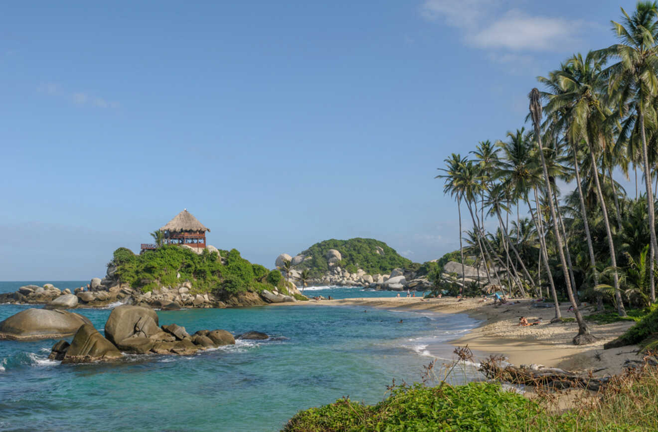A small island with a thatched-roof structure is surrounded by clear blue water and rocky shores. A sandy beach with palm trees lines the right edge of the image under a blue sky with few clouds.