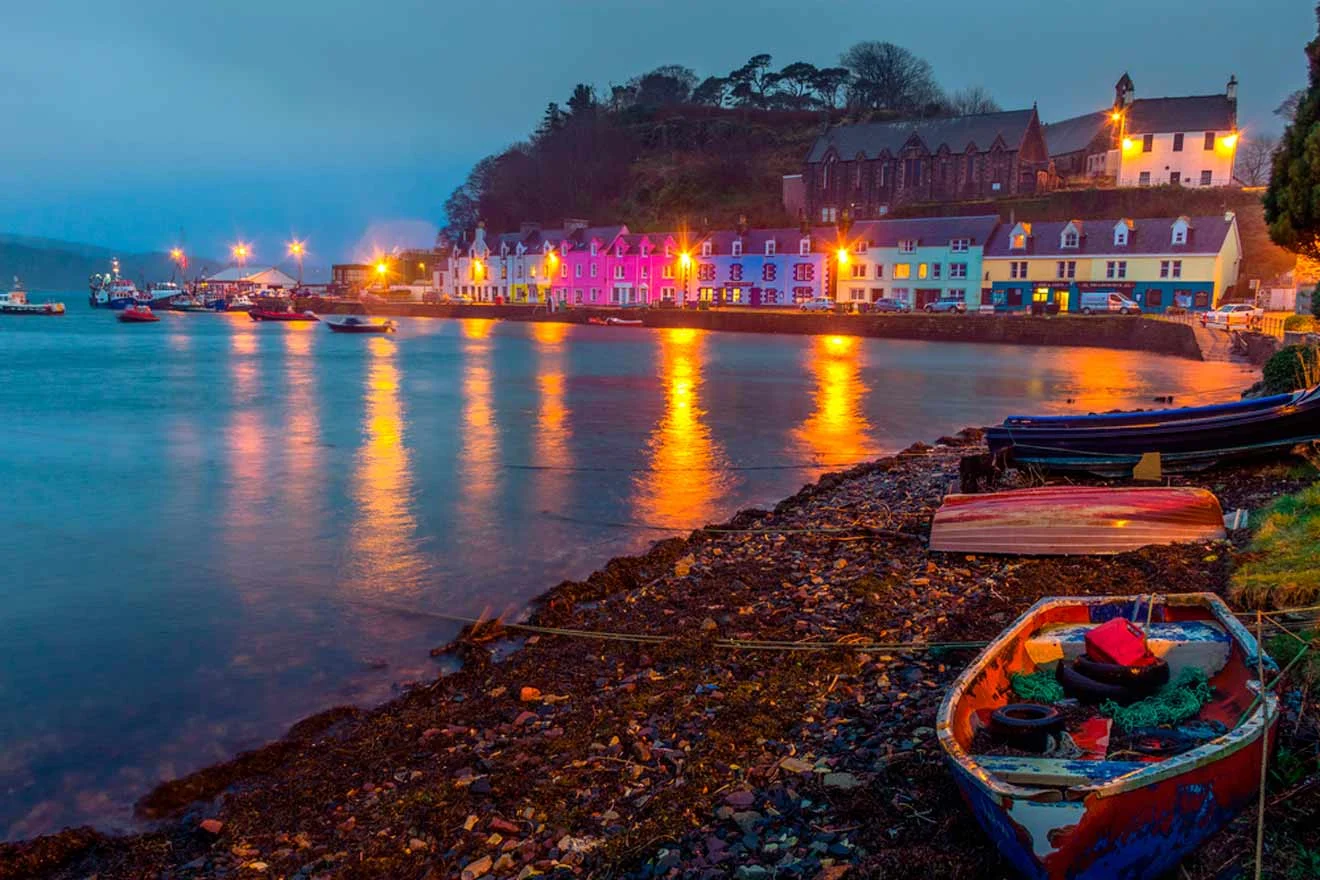 Colorful houses line the shore of a calm coastal village at dusk, with reflections in the water and boats resting on the rocky beach.