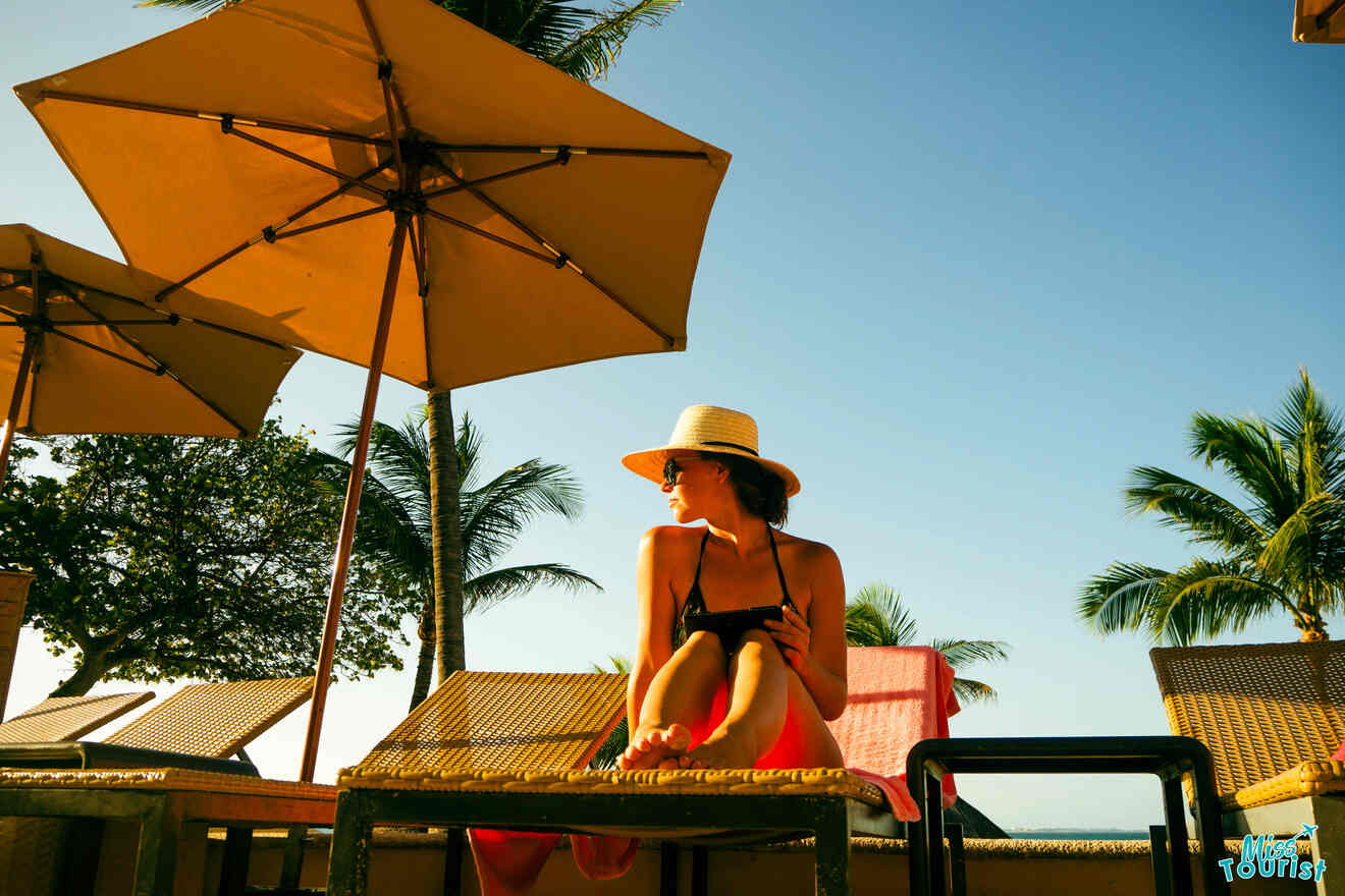 the author of the posts in a sun hat and swimsuit sits on a lounge chair under an umbrella, with palm trees and a clear sky in the background.