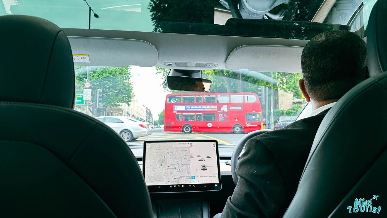 View from the backseat of a car showing the driver and a navigation screen, with a red double-decker bus visible through the windshield in London traffic.