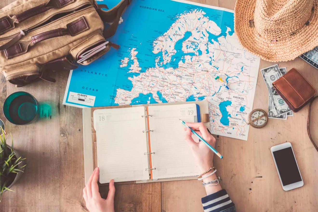 A person writing in a planner on a table with a map of Europe, a brown bag, a wristwatch, a smartphone, a green drink, and a straw hat.