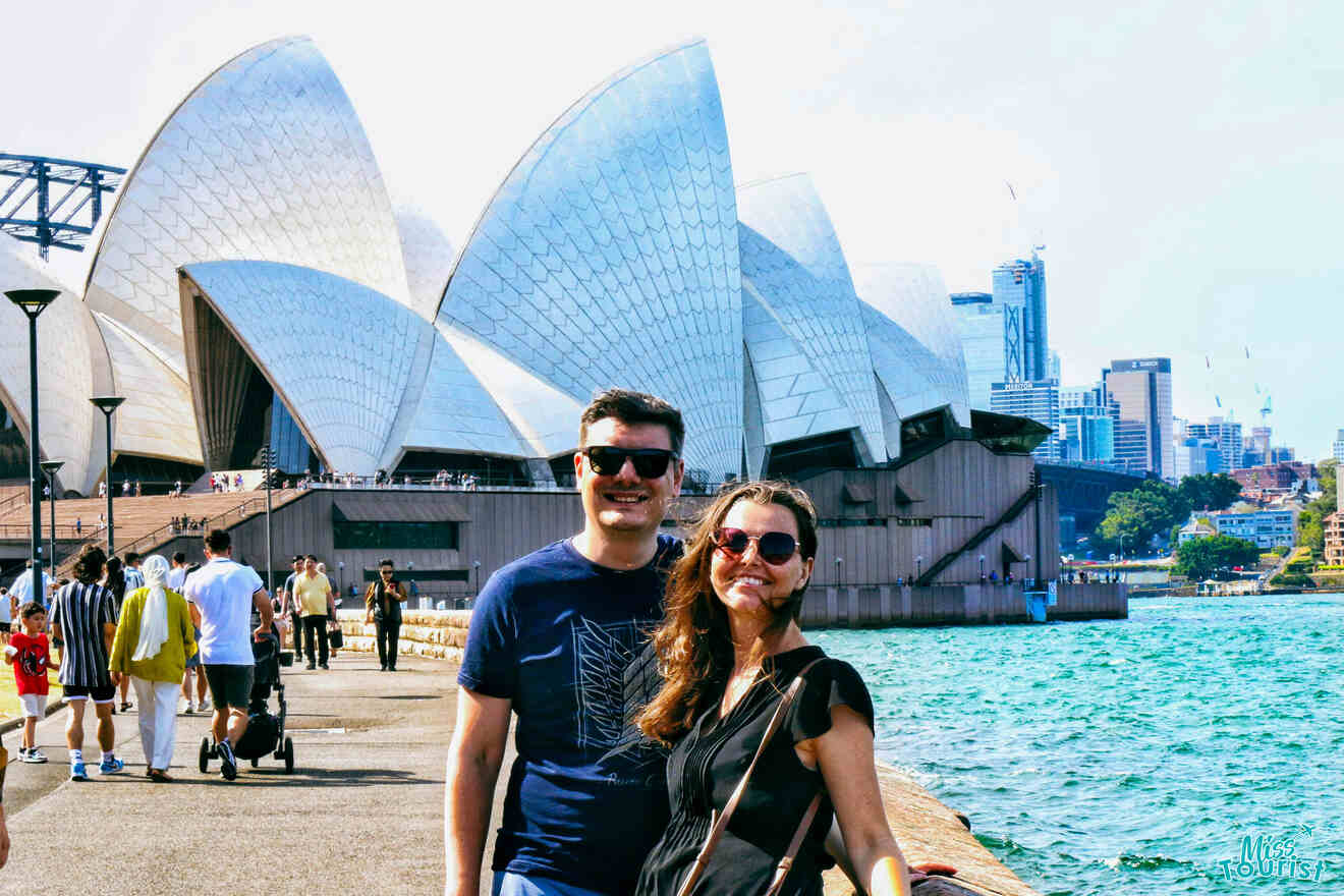 A smiling man and woman stand in front of the Sydney Opera House, with people walking around and the city skyline visible in the background.