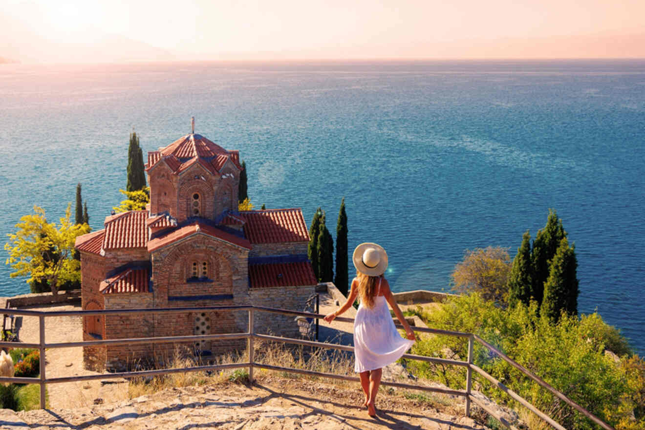 A person in a white dress and sun hat stands on a viewpoint overlooking a historic building with a red-tiled roof and a large body of water in the background.
