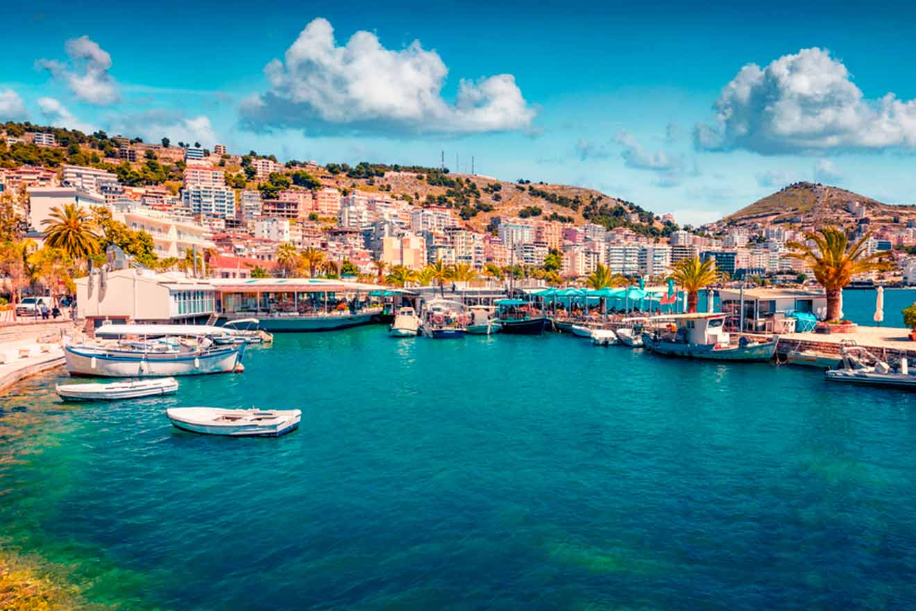 Scenic view of a coastal town with numerous buildings on a hillside, palm trees lining the waterfront, and boats docked in the clear blue waters of a harbor under a partly cloudy sky.