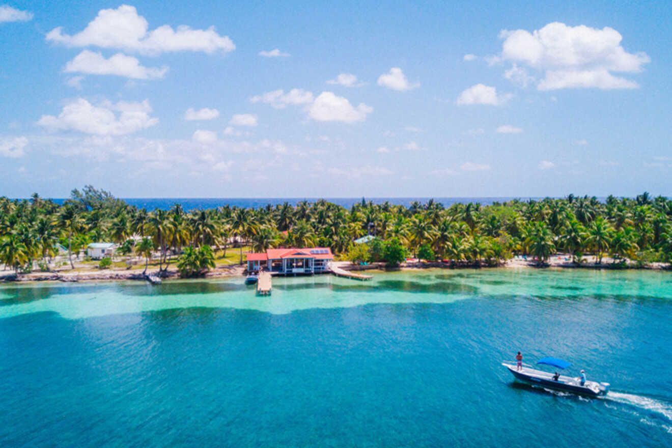 A boat navigates clear blue water near a tropical island with palm trees and a few buildings along the shoreline under a partly cloudy sky.