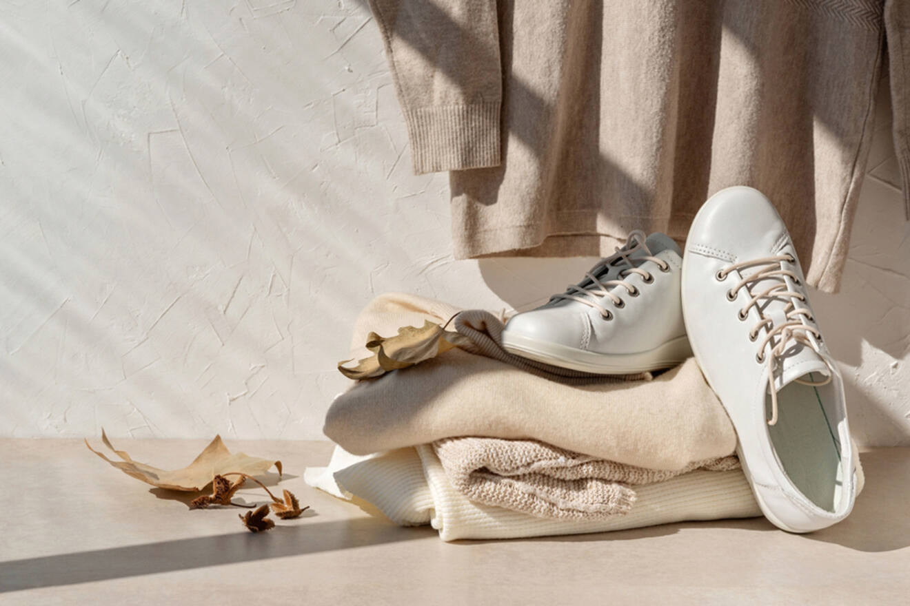 White sneakers placed on a stack of beige and cream-colored folded clothes with dried leaves scattered around, against a textured white wall with a sweater hanging above.