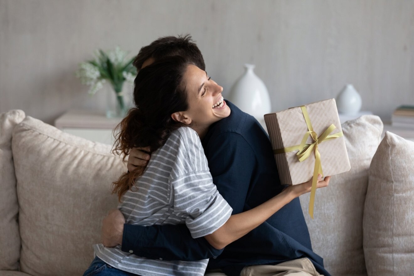 A person with curly hair smiles while hugging another person on a couch, holding a wrapped gift with a yellow ribbon behind their back. The background includes a vase with flowers and a book on a table.