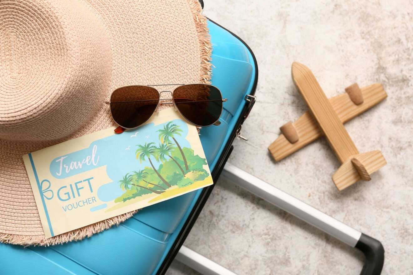 A blue suitcase with a beige hat, a pair of sunglasses, a travel gift voucher, and a small wooden airplane on a light-colored surface.