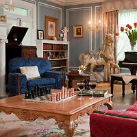 A well-decorated living room with blue walls features a chessboard on a wooden coffee table, a blue armchair, bookshelves, a grand piano, and a horse figurine by a window with curtains.