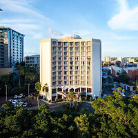 A tall, modern hotel building with multiple floors is situated among trees and urban structures under a clear sky.