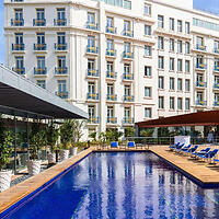 A modern hotel exterior with multiple stories and balconies overlooks a rectangular outdoor pool with blue lounge chairs arranged around the poolside.