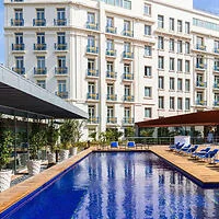 A modern hotel exterior with multiple stories and balconies overlooks a rectangular outdoor pool with blue lounge chairs arranged around the poolside.
