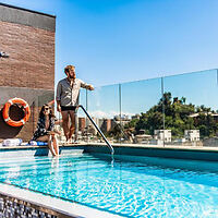 A couple enjoying a sunny day by a rooftop swimming pool with a clear blue sky and cityscape in the background.