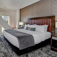 A modern hotel room with a king-sized bed, white linens, a gray throw blanket, and patterned wallpaper. The bedside tables hold lamps, and a window allows natural light to flood the room.