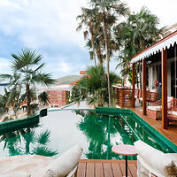 A tropical outdoor patio with a rectangular pool, surrounded by palm trees and lounge chairs, adjacent to a building with a covered veranda.