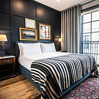 A well-decorated bedroom with a large bed, striped bedding, two bedside tables with lamps, framed pictures on a dark accent wall, and floor-to-ceiling curtains next to a large window.