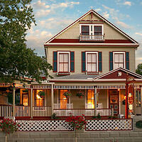 A two-story house with a wraparound porch, white and red trim, large windows, and lit interior lights. A tree is partially visible on the left side.