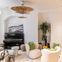 A modern living room featuring a black piano, white cushioned chairs with green pillows, a small round table, and a large indoor plant. Minimalist decor with a neutral color palette.