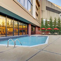 Outdoor swimming pool next to a building with glass windows, surrounded by a concrete deck with tables and red umbrellas. Pine trees partially visible in the background.