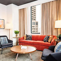 A modern living room with a red sofa, two black chairs, a round wooden coffee table, floor lamp, indoor plants, and a large window showcasing a cityscape outside.