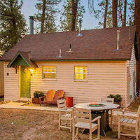 A small, quaint house with a green front door, illuminated by outdoor lights at dusk. The yard features wooden patio furniture, including a round table and chairs. Tall trees stand in the background.