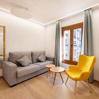 A minimalist living room with a grey sofa, two small round wooden tables, a yellow armchair, and a window with green curtains.