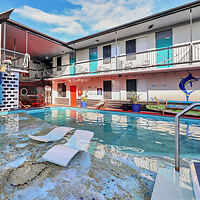 An outdoor swimming pool area with two submerged lounge chairs. The building in the background has multiple doors painted in various colors, a mural of a fish, and an upper balcony with a railing.