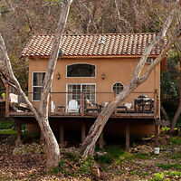 A small beige house with a tiled roof and a wooden deck, surrounded by tall trees and situated in a wooded area.