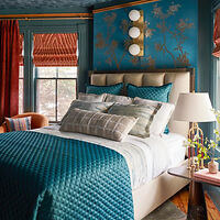 A bedroom with teal walls and a patterned wallpaper accent. The bed has a teal quilt and multiple pillows. There are red curtains, two windows with woven blinds, and a lamp on a nightstand.