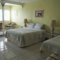 A bedroom with two double beds, lamps on bedside tables, a small dining area by the window, and light-colored walls with a framed picture.