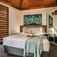 A neatly made bed with a hat and green throw on the foot, flanked by a nightstand and mirrored closet door. Above the bed is a tropical-themed artwork. Wooden ceiling and louvered window on the left.