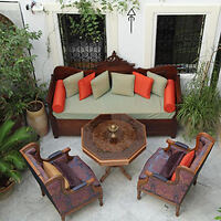 A wooden outdoor seating area with a green cushioned couch and red pillows, two armchairs, and a hexagonal wooden table. The space is decorated with plants and set on a light-colored patio.