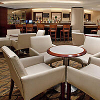 A modern lounge area with white upholstered chairs around circular tables in the foreground and a bar counter with wooden stools in the background.