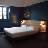 A hotel room with a double bed against a dark blue wall, lit by two bedside lamps. There is a window to the left and a wooden bedside table with a phone on the right.