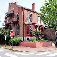 A two-story red brick building with red trim, a fire escape on the side, and a "Do Not Enter" sign on the corner. There are trees and bushes around the building.