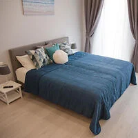 A neatly made bed with blue bedding, pillows, and a headboard. There are bedside tables with lamps on both sides, and the room has light-colored walls and floor, with curtains partially open.