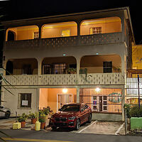 A two-story building at night with parked red SUV in front. Upper and lower floors have balcony areas, and there are potted plants beside the entrance on the ground floor.