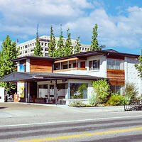 A modern two-story building with large windows, wood and white exterior, and greenery, situated alongside a road with a taller building in the background under a partially cloudy sky.