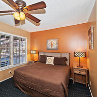 A bedroom with an orange accent wall, a bed with brown bedding, two bedside tables with lamps, a ceiling fan, and a window with white blinds.